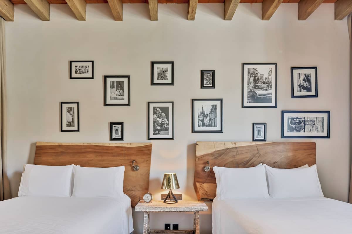 Quarto de hotel com duas camas, uma mesinha com abajur e parede decorada com fotografias em preto e branco.