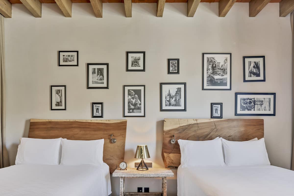A hotel room with two beds, a small table with a lamp, and a wall adorned with framed black and white photographs.
