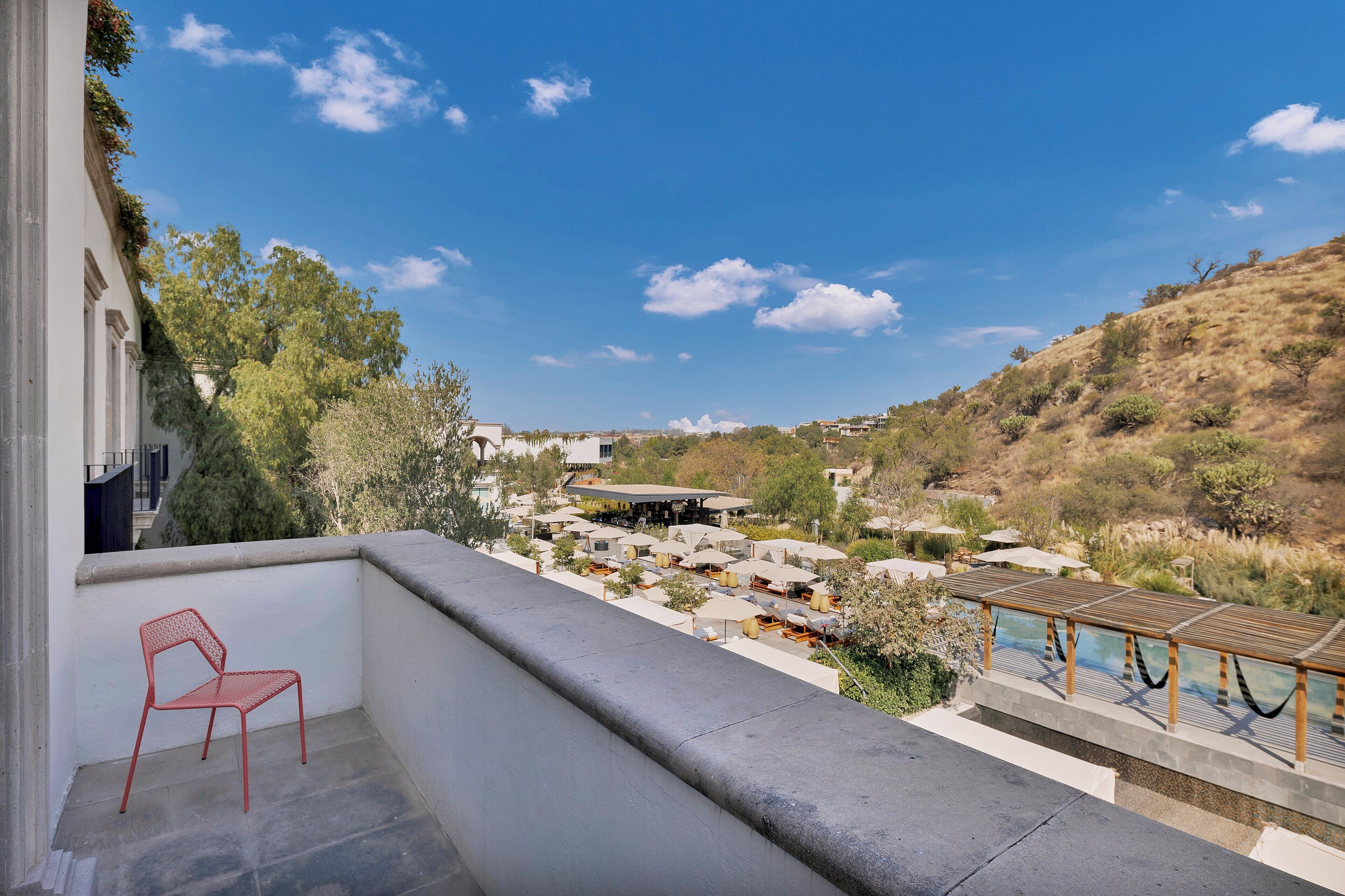 A balcony with a red chair, a pool area, and a view of a resort with multiple buildings and a hilly landscape.