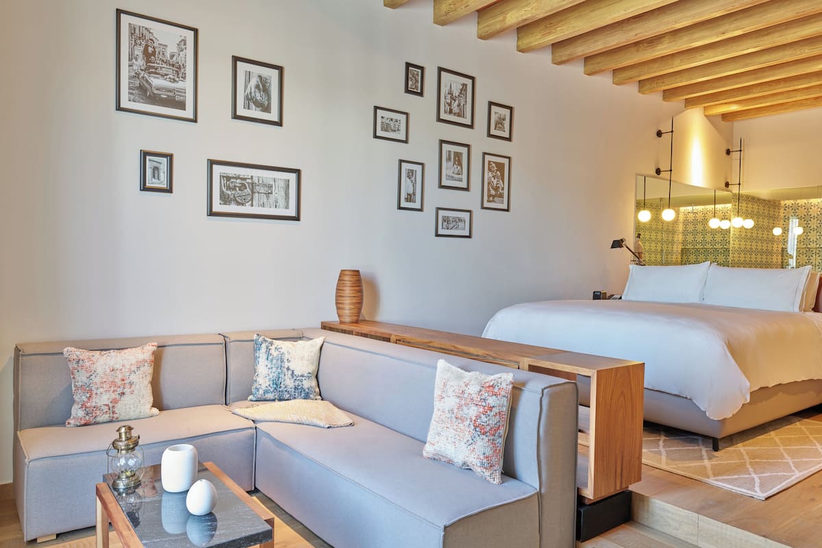 Quarto de hotel moderno com sofá cinza claro, um banco de madeira, mesa de centro de vidro e cama com roupa de cama branca.