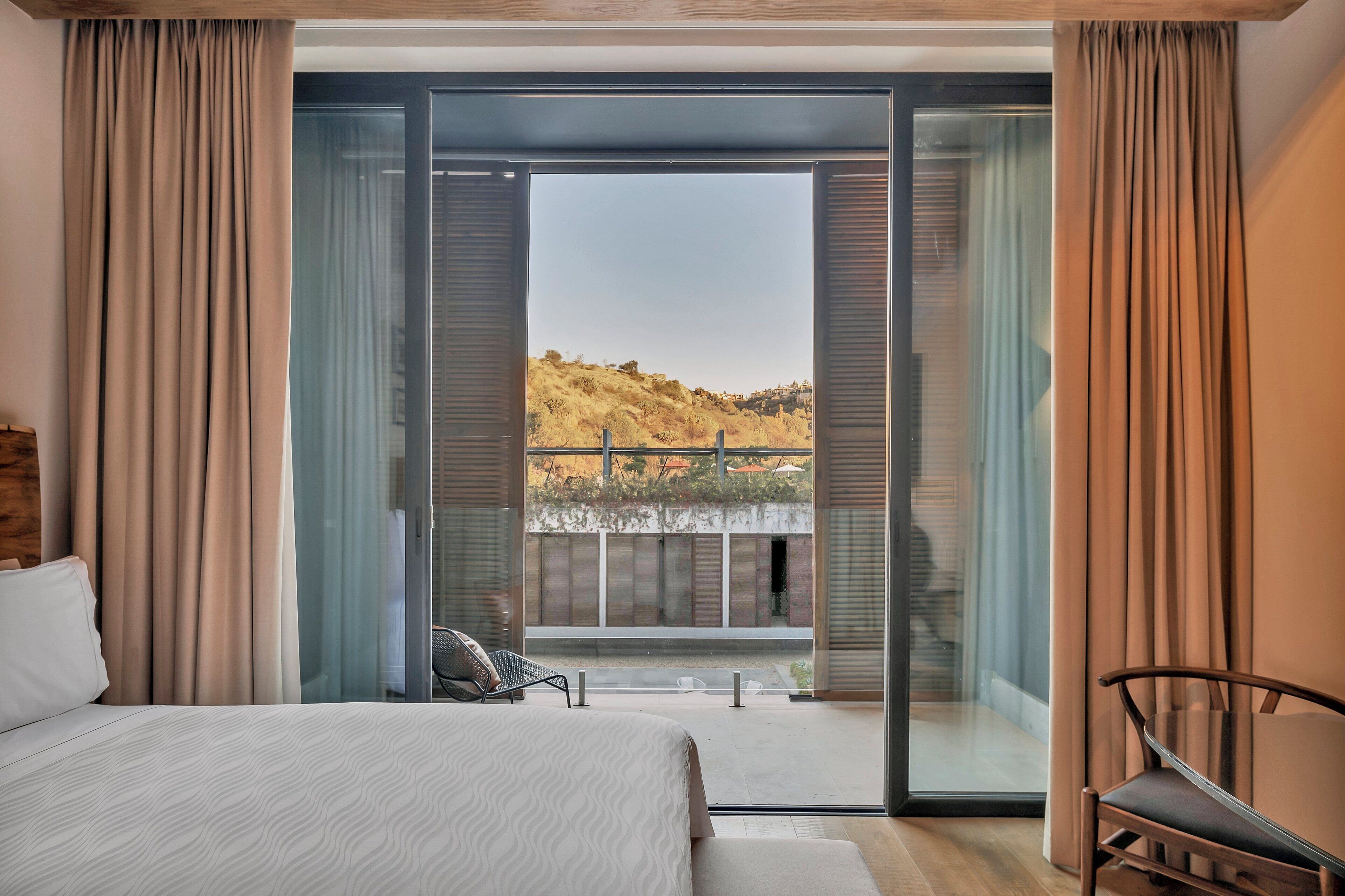 A bedroom with a large bed, a chair, and a sliding glass door leading to a balcony with a view of hills.