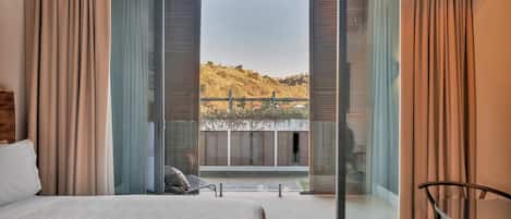 Un dormitorio con una cama grande, una silla y un ventanal corredero que da a un balcón con vistas a las colinas.