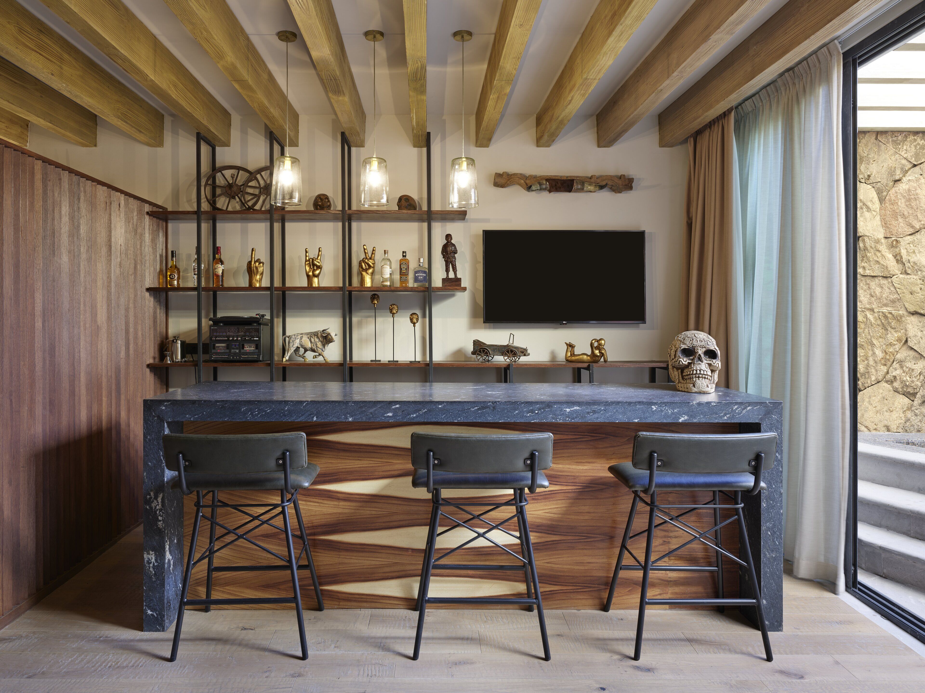 A modern kitchen with a marble countertop, three bar stools, wooden beams, and a large TV.