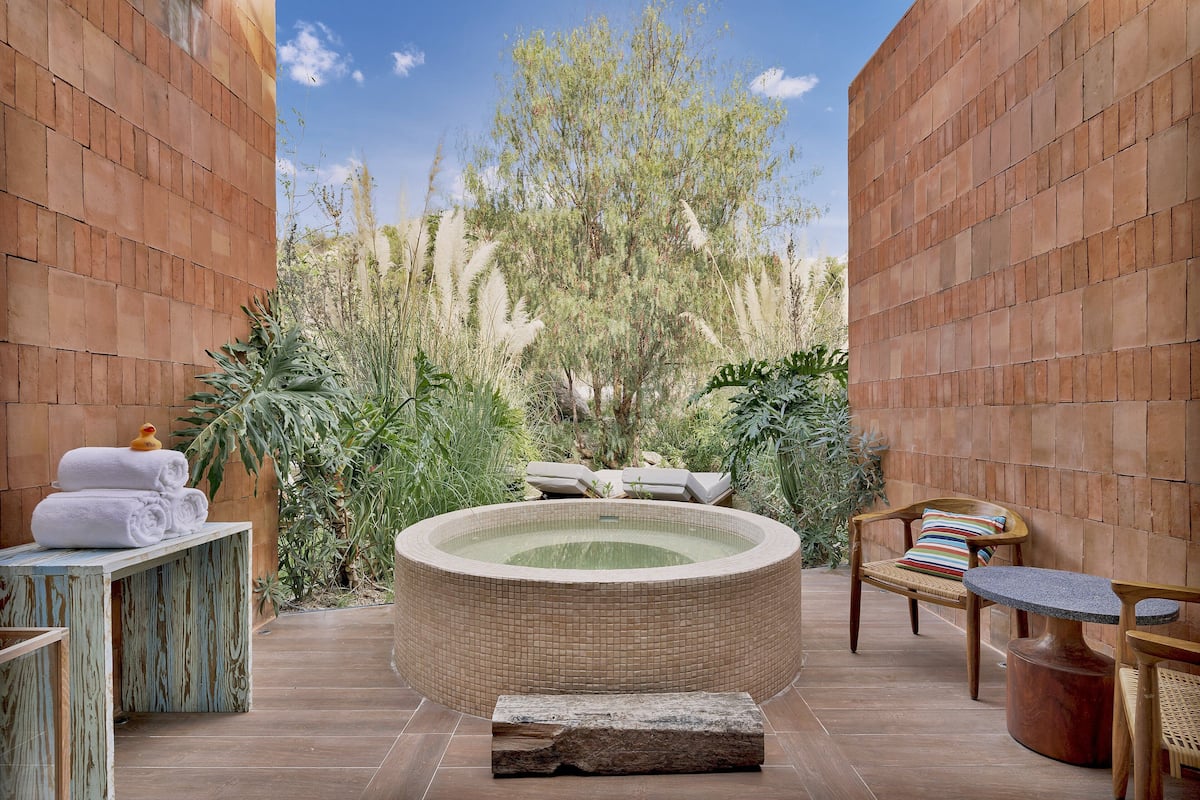 A hot tub surrounded by wooden benches and a table with towels, set against a brick wall with a view of trees and plants.