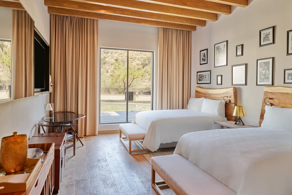 Quarto de hotel com duas camas, uma escrivaninha, uma cadeira, uma televisão e uma janela grande com cortinas.