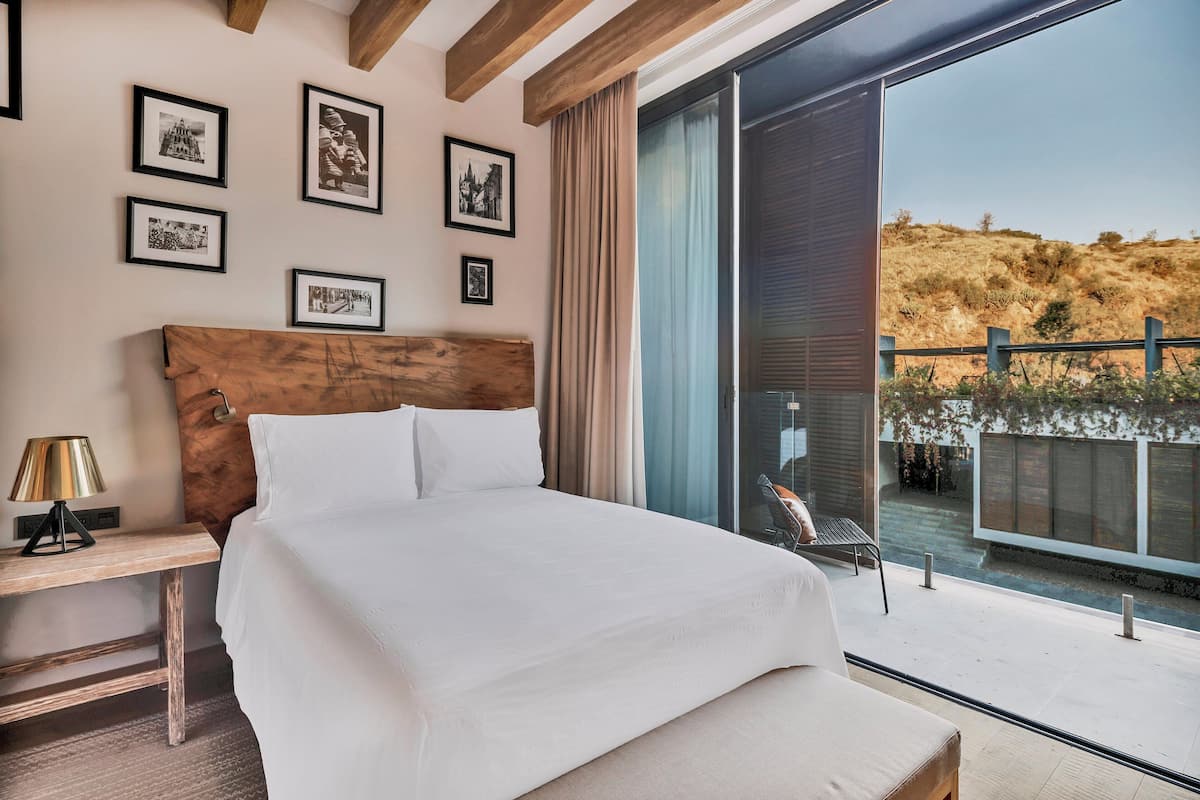 Quarto com cama grande, mesa de cabeceira, luminária e vista para uma encosta através de uma porta de vidro deslizante.