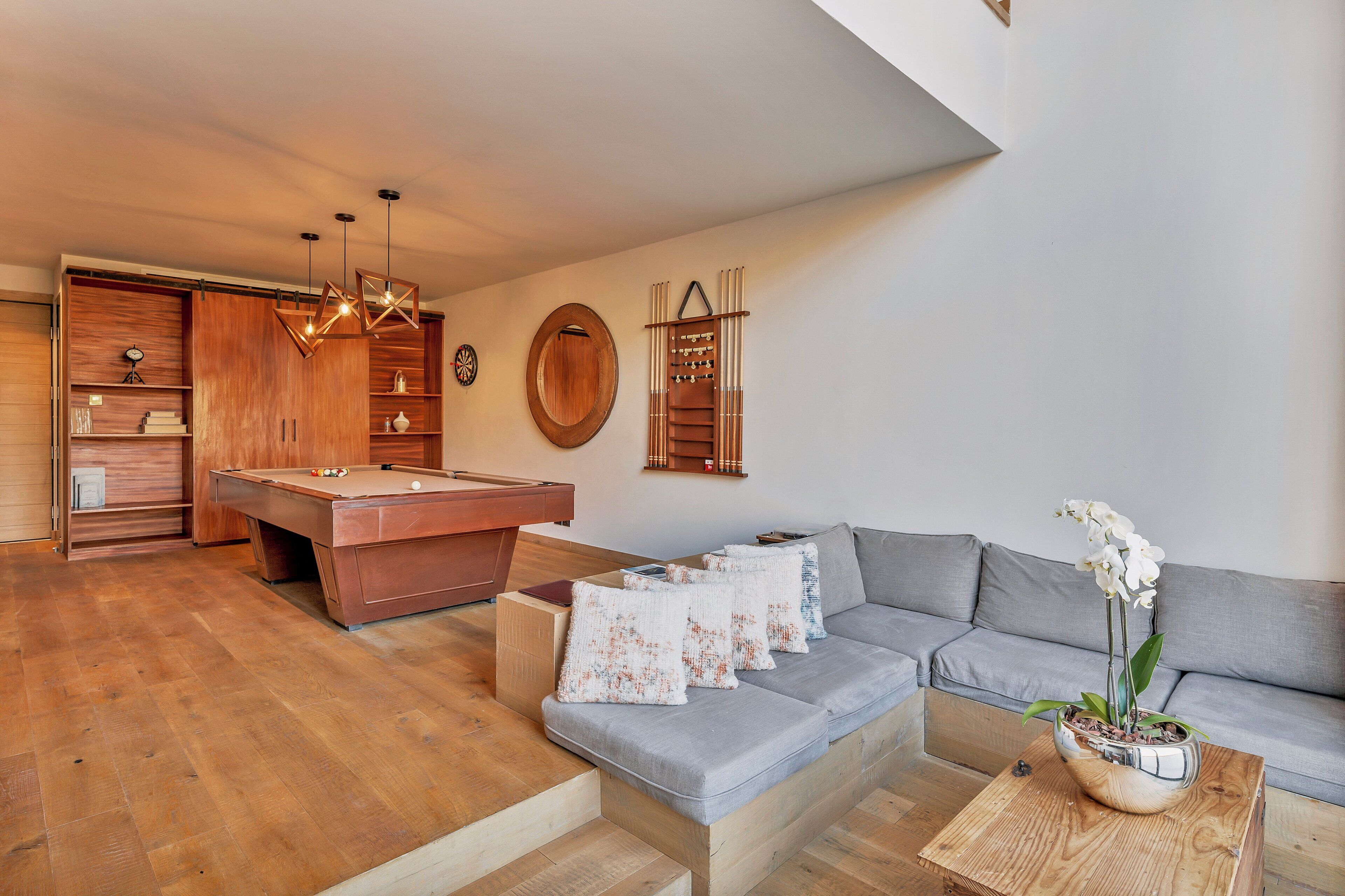 A modern living room with a pool table, grey sofa, wooden cabinets, and a potted orchid.