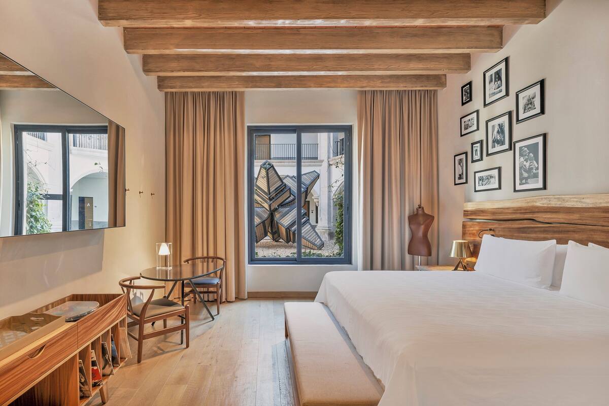Quarto de hotel com uma cama grande, uma escrivaninha com cadeira, uma janela com cortinas e um painel com quadros.