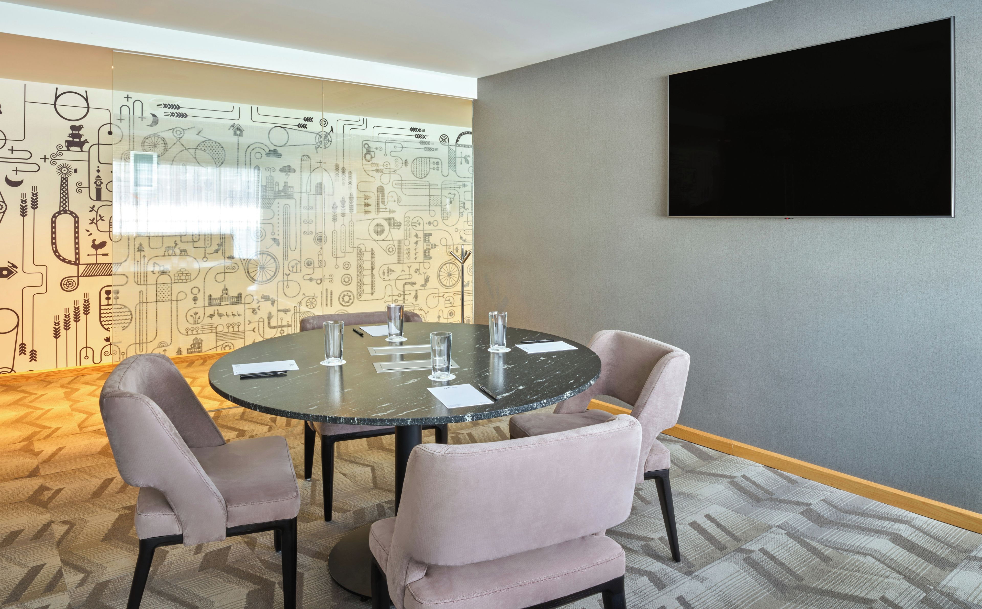A conference room with a round table, four chairs, a flat-screen TV on the wall, and patterned wallpaper.