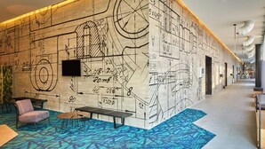 A hotel lobby with a large wall of architectural drawings, a seating area with a patterned carpet, and a long corridor with a modern design.