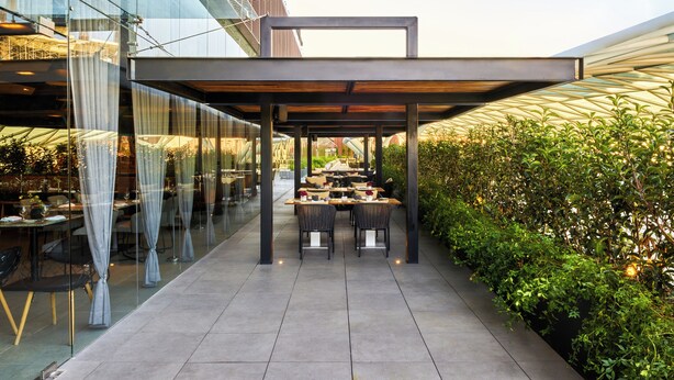 An outdoor dining area with tables and chairs, a glass wall, and a metal structure overhead.