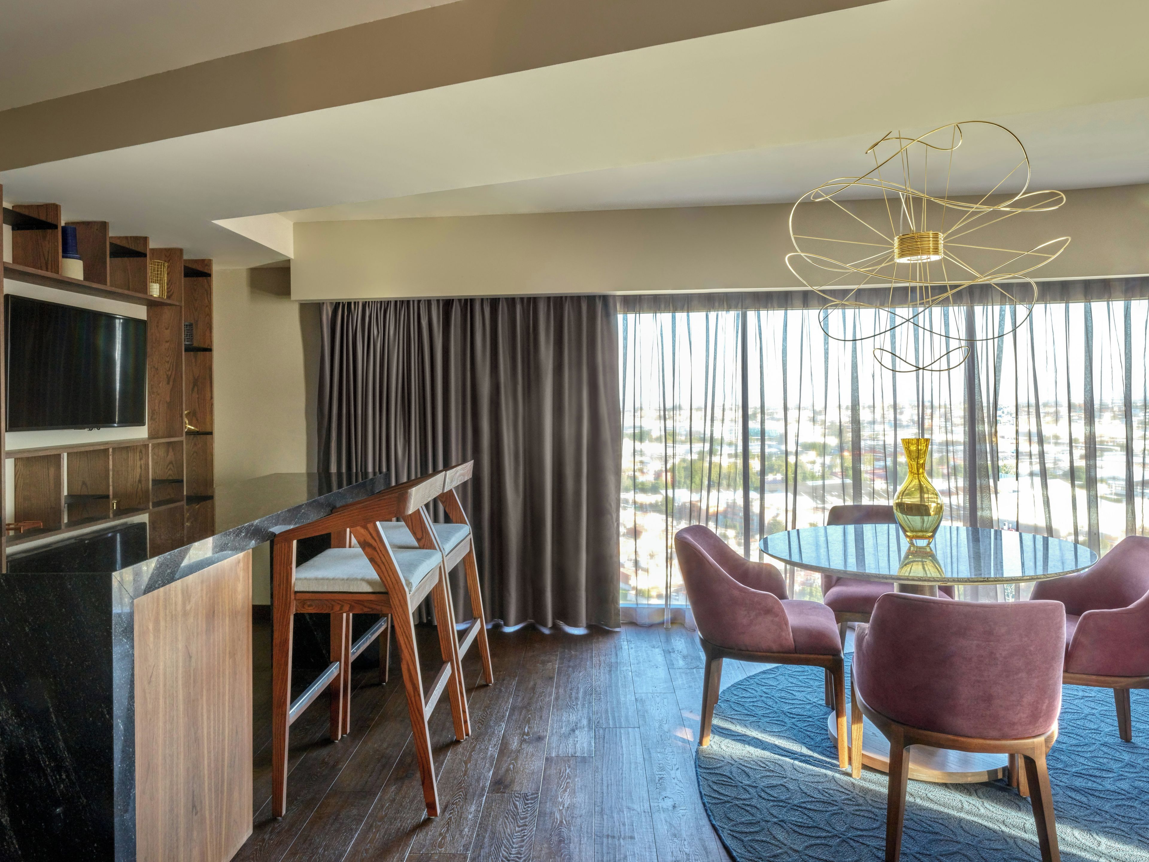 A modern hotel room with a dining area, a TV mounted on a wooden shelf, and a view of the city through large windows.