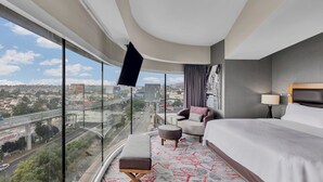 A modern hotel room with a large window offering a city view, a bed, a seating area with a sofa and ottoman, and a flat-screen TV mounted on the wall.