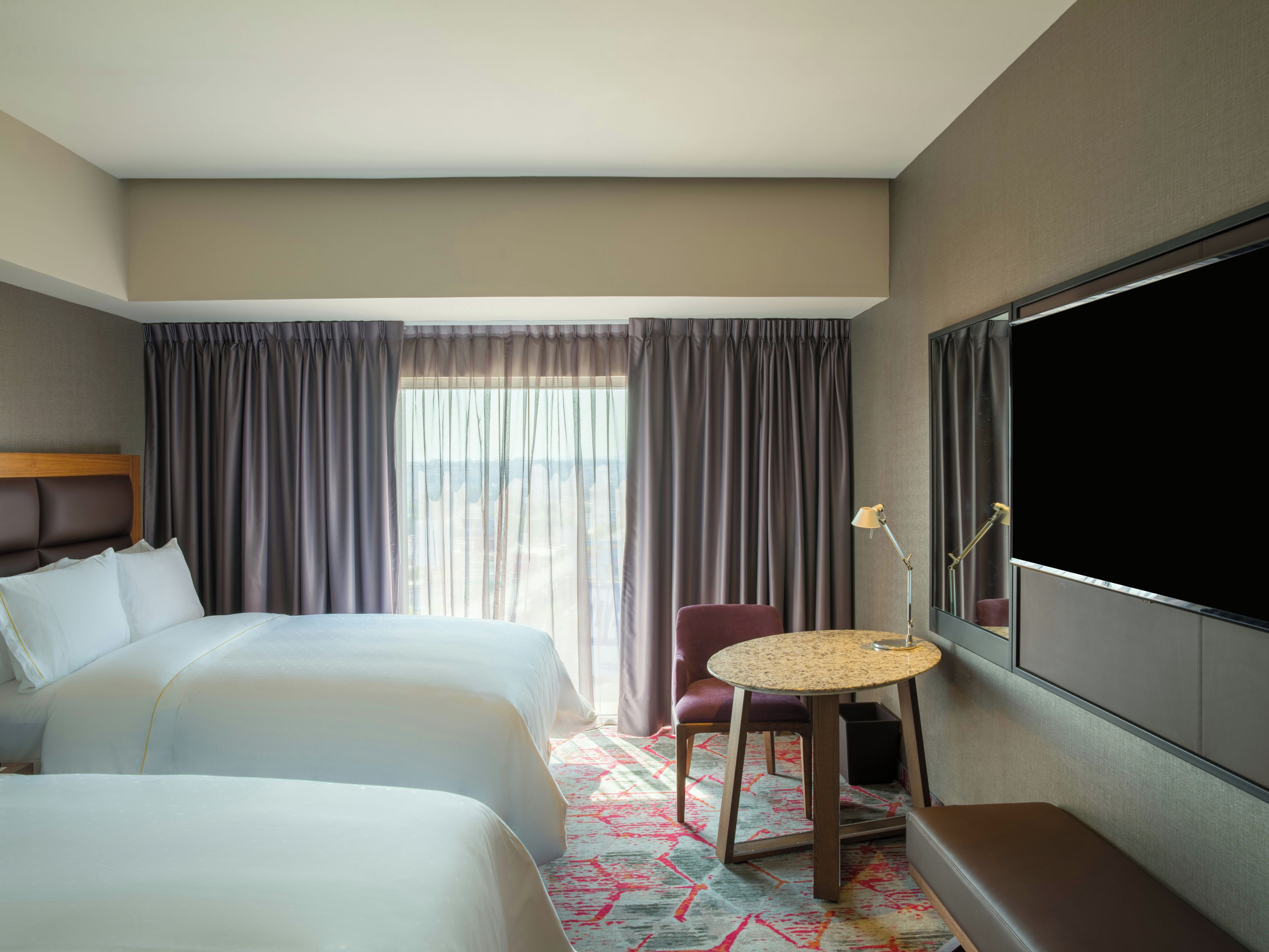 Une chambre d’hôtel avec deux lits, un coin salon avec une table, une grande fenêtre avec des rideaux et un téléviseur à écran plat.