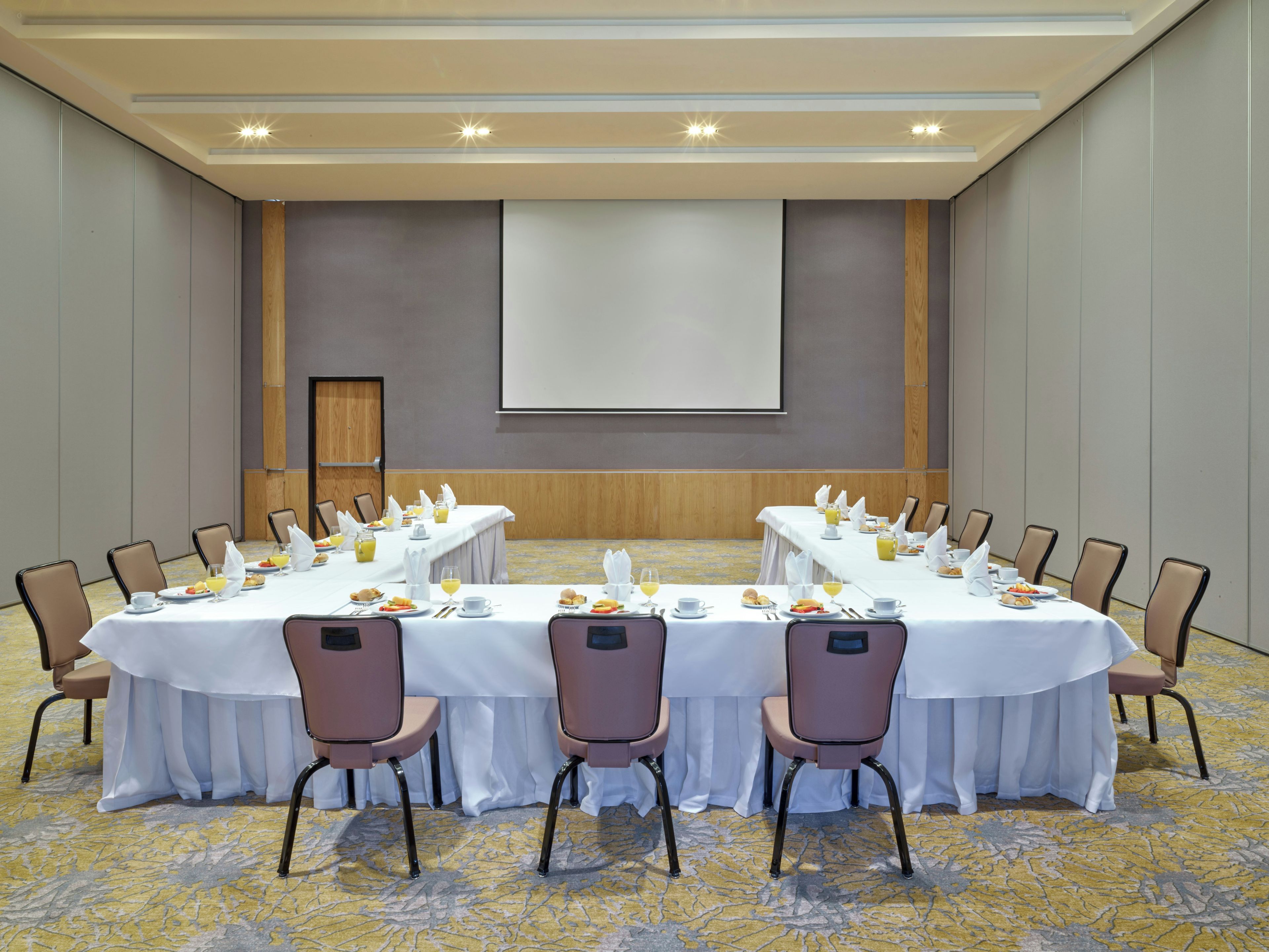 Un salón de conferencias con una mesa larga, sillas y una pantalla de proyector.
