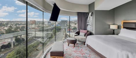 A hotel room with a large window, a bed, a chair, a small table, and a view of a cityscape.