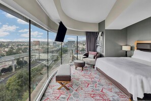A hotel room with a large window, a bed, a chair, a small table, and a view of a cityscape. - Fiesta Americana México Satélite (Tlalnepantla de Baz)