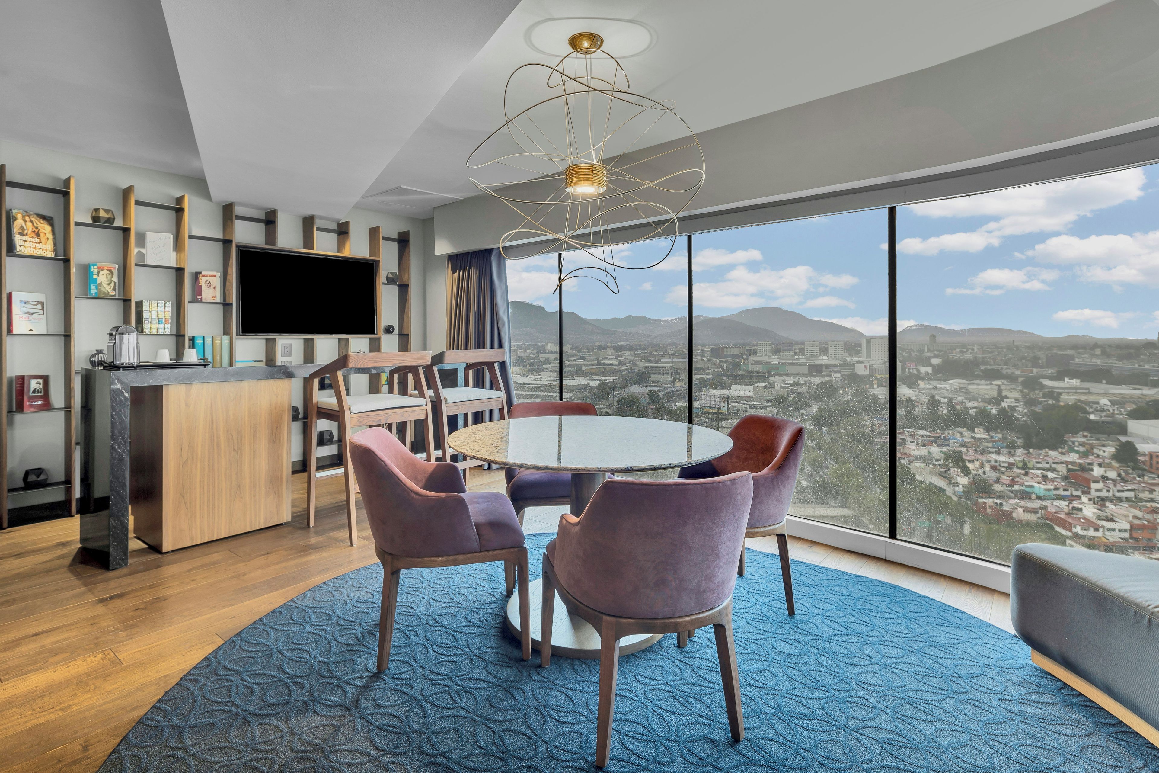 A modern living room with a round table, purple chairs, a large window with a city view, a flat-screen TV, and a unique ceiling light fixture.