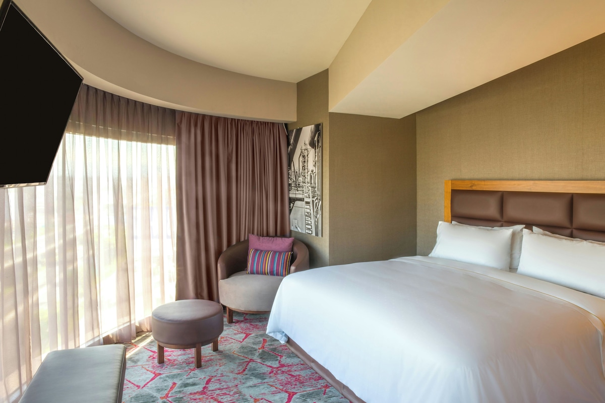 A hotel room with a large bed, a chair with a pillow, a small ottoman, a wall-mounted TV, and a patterned rug.