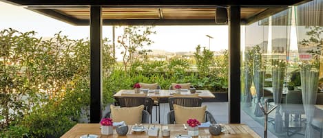 Un espacio de comedor al aire libre con mesa y sillas de madera, rodeado de vegetación y vistas panorámicas de la ciudad.