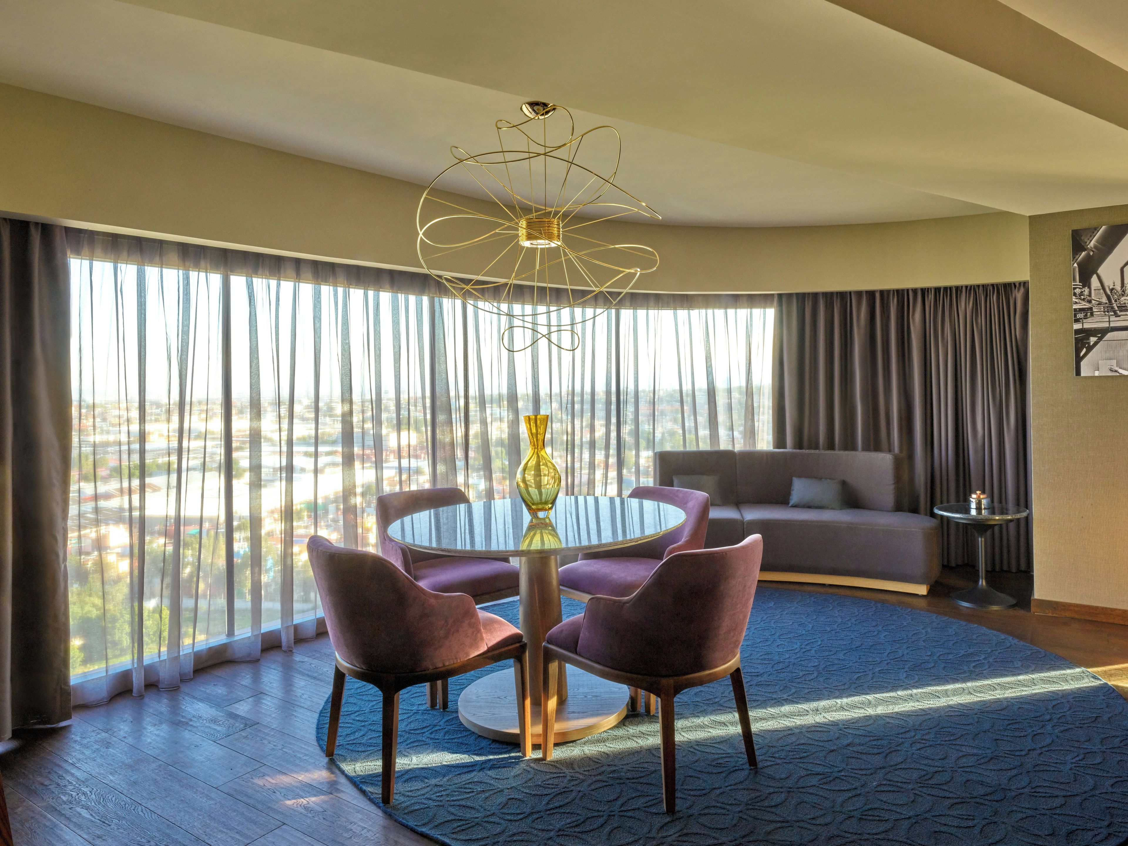 Habitación de hotel con una mesa redonda, dos sillas, un sofá, una mesita auxiliar y vistas de la ciudad a través de grandes ventanales.