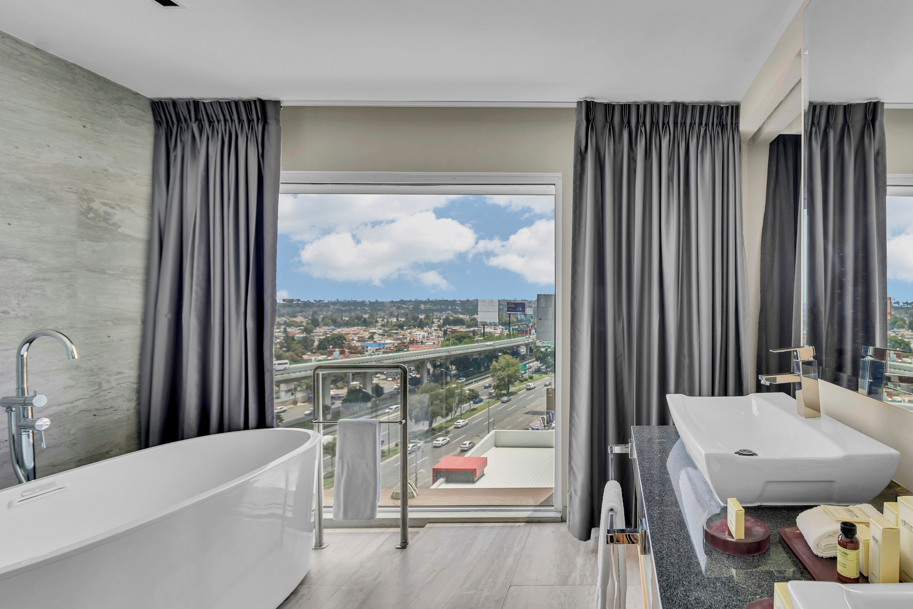 Un baño moderno con bañera, un lavamanos amplio y vistas a un paisaje urbano a través de un ventanal grande.