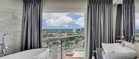 Un baño moderno con bañera, un lavamanos amplio y vistas a un paisaje urbano a través de un ventanal grande.