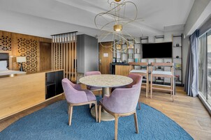 Una habitación de hotel moderna con una mesa de comedor redonda, sillas moradas, alfombra azul, televisor y un panel de madera con un diseño geométrico.
