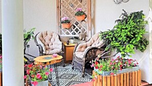 Un rincón al aire libre acogedor con sillas acolchadas y una pequeña mesa, rodeado de plantas en macetas y un adorno de pared decorativo.