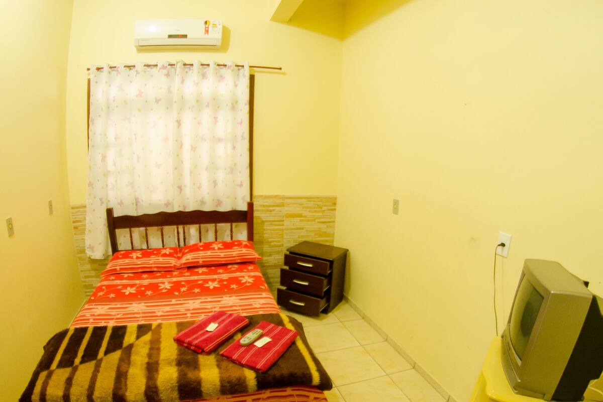 Une petite pièce, bien éclairée, avec un lit, un téléviseur posé sur un meuble et une petite table.