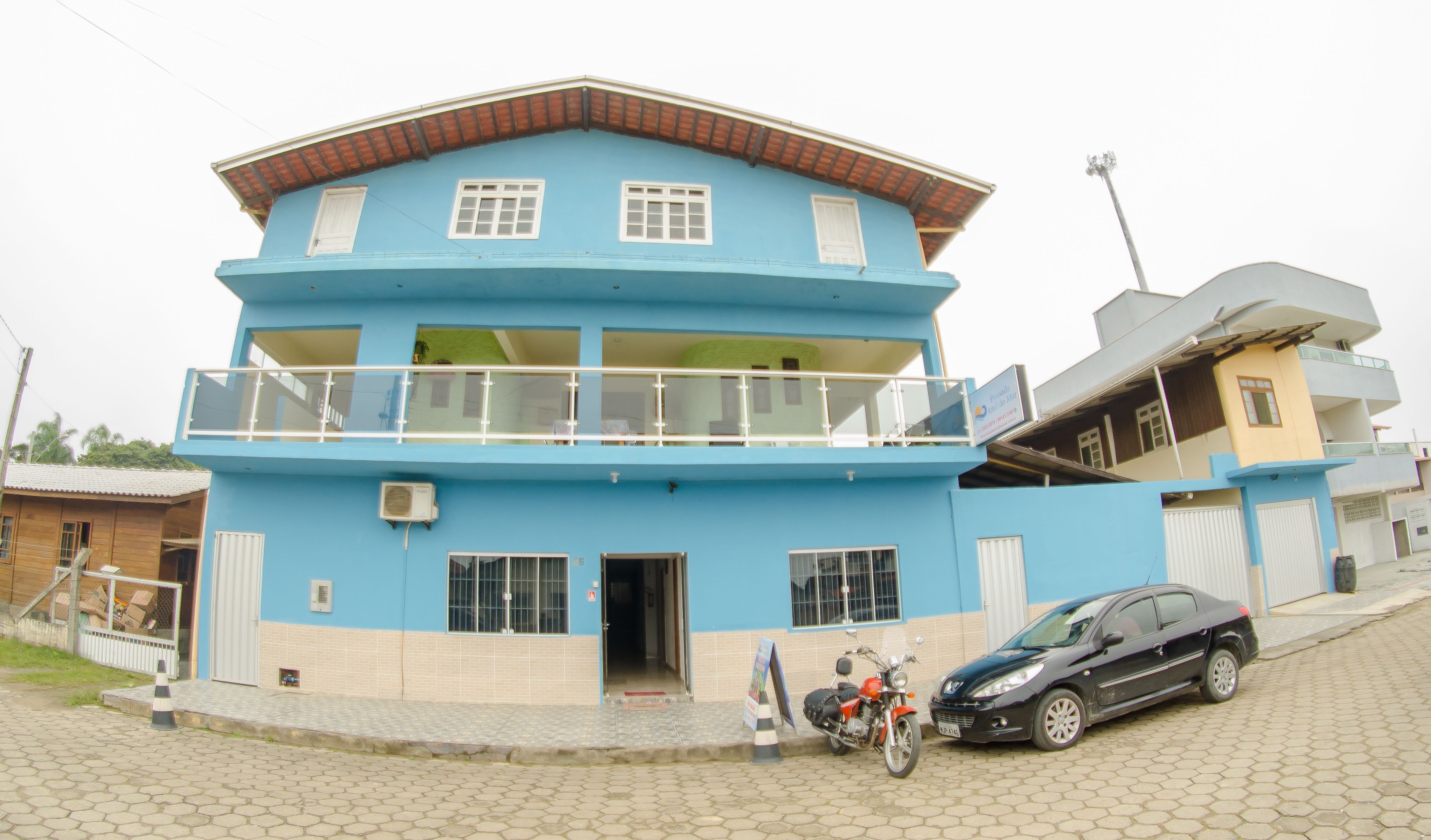 A multi-story building with a blue facade, a balcony on the upper floor, and a parked car and motorcycle in front.