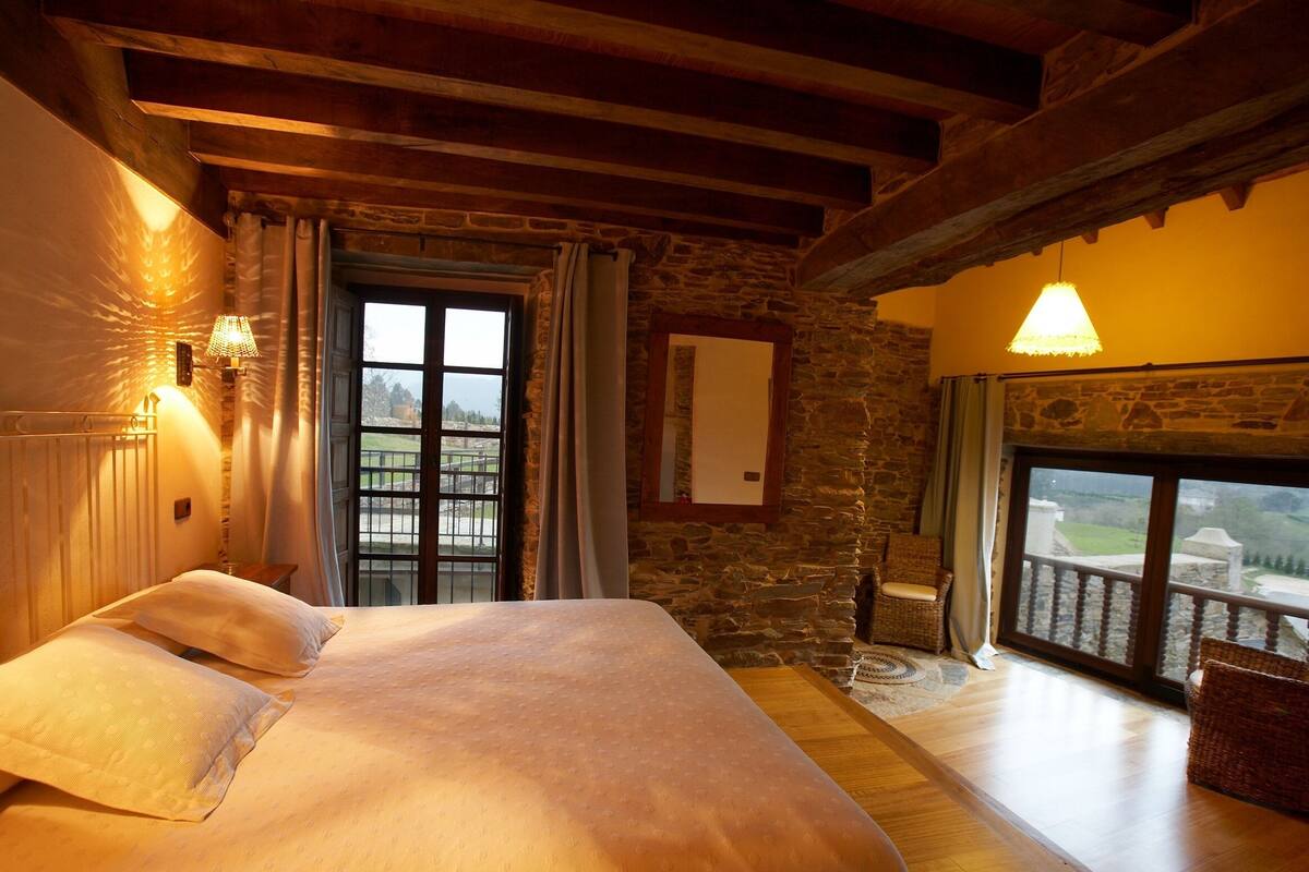 Une chambre avec un grand lit, des murs en pierre, des poutres en bois, un balcon avec des fauteuils en osier et une vue sur la campagne.