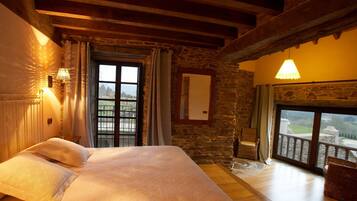 A bedroom with a large bed, stone walls, wooden beams, a balcony with wicker chairs, and a view of the countryside.