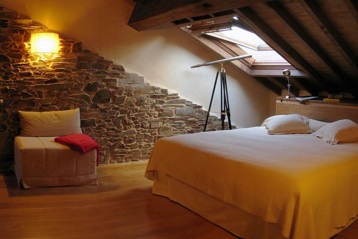 Une chambre confortable avec un mur en pierre, un plafond en bois, un lit avec une literie blanche et une lucarne.