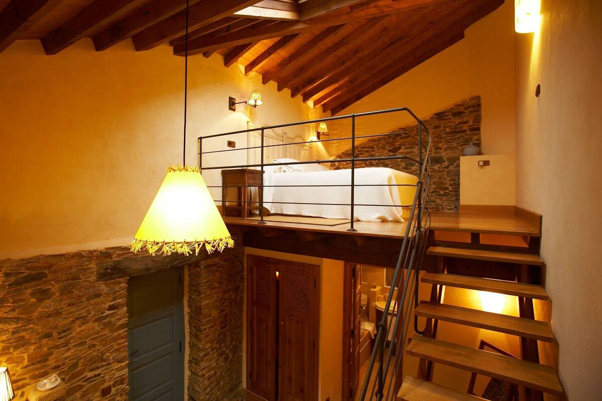 Une chambre confortable située dans les combles, avec un escalier en bois, une cheminée en pierre et un lit doté d’une balustrade en métal.