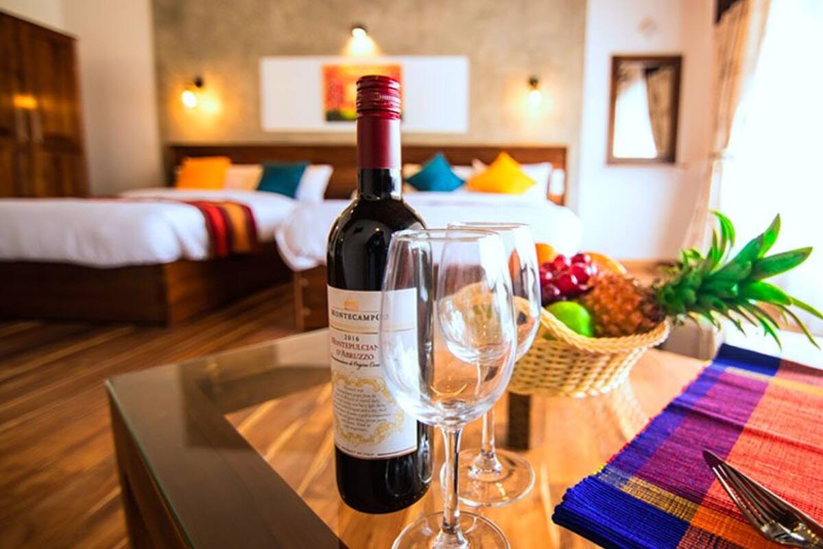 A hotel room with a bed, a bedside table with a bottle of wine and glasses, a fruit basket, and a colorful table runner.
