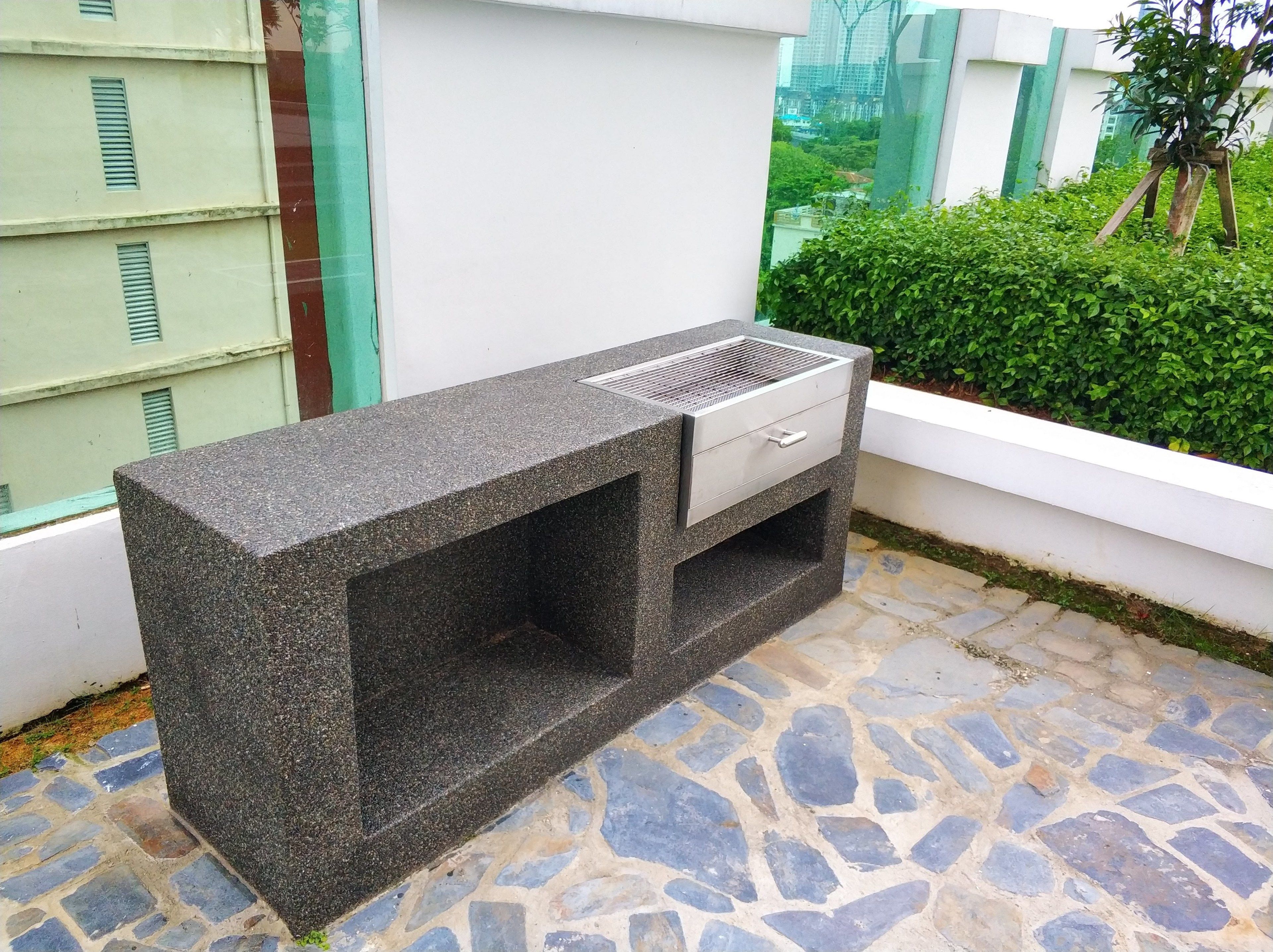 Une cuisine extérieure en granit, dotée d’un barbecue intégré, située sur une terrasse dallée de pierres, à côté d’un mur blanc et d’une haie verte.