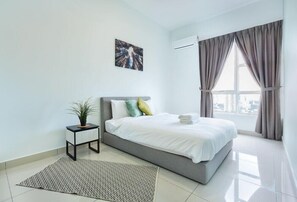 A modern bedroom with a grey bed, white bedding, a small bedside table with a plant, and a large window with curtains.