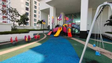 A playground with slides and swings, surrounded by a building with balconies and a pool area.