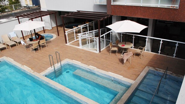 A rooftop swimming pool with lounge chairs and umbrellas, surrounded by a wooden deck and a glass railing.