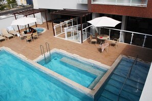 Zwembad op een dak met ligbedden en parasols, omringd door een houten terras en een glazen balustrade.