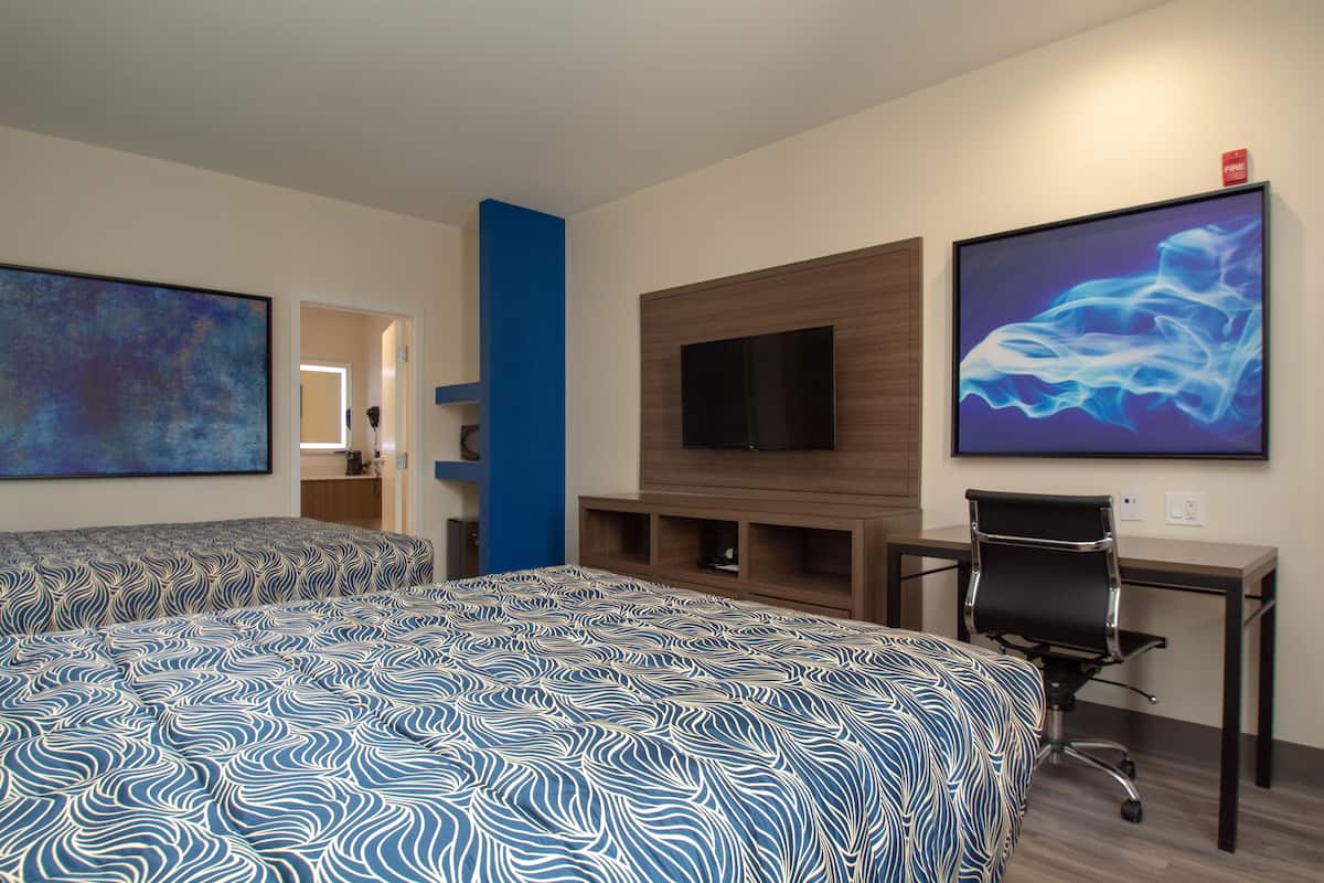 Quarto de hotel com duas camas, uma escrivaninha, uma cadeira, televisão e obras de arte nas paredes.
