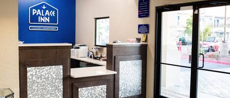 Zona de recepción con un letrero azul que dice "PALACE INN LUXURY Redefined", un mostrador de recepción, una puerta de cristal con un letrero y un felpudo azul con la inscripción "NNN".
