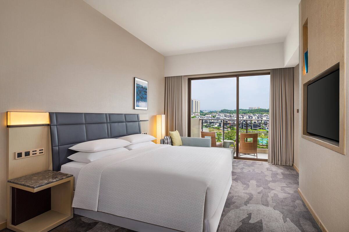Quarto de hotel com cama grande, mesa de cabeceira, televisão e varanda com vista para a cidade.