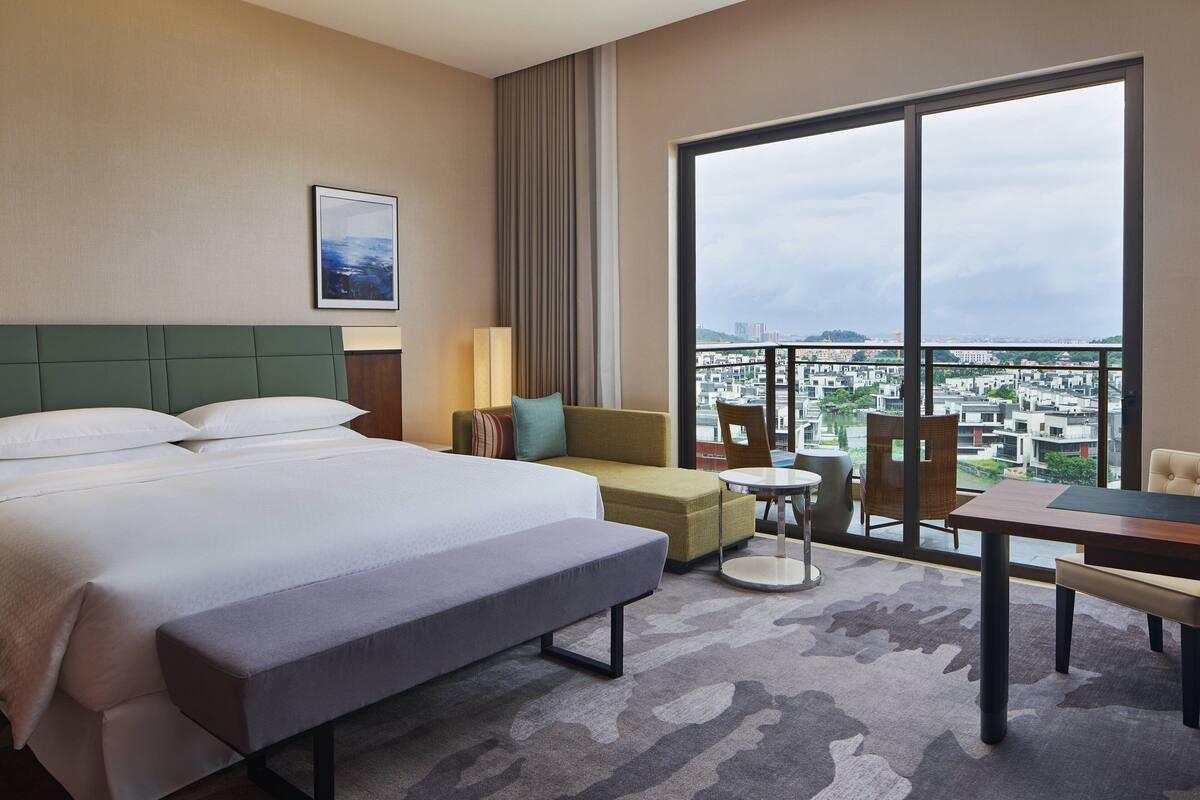 Quarto de hotel com uma cama grande, um sofá, uma mesinha e varanda com vista para a cidade.