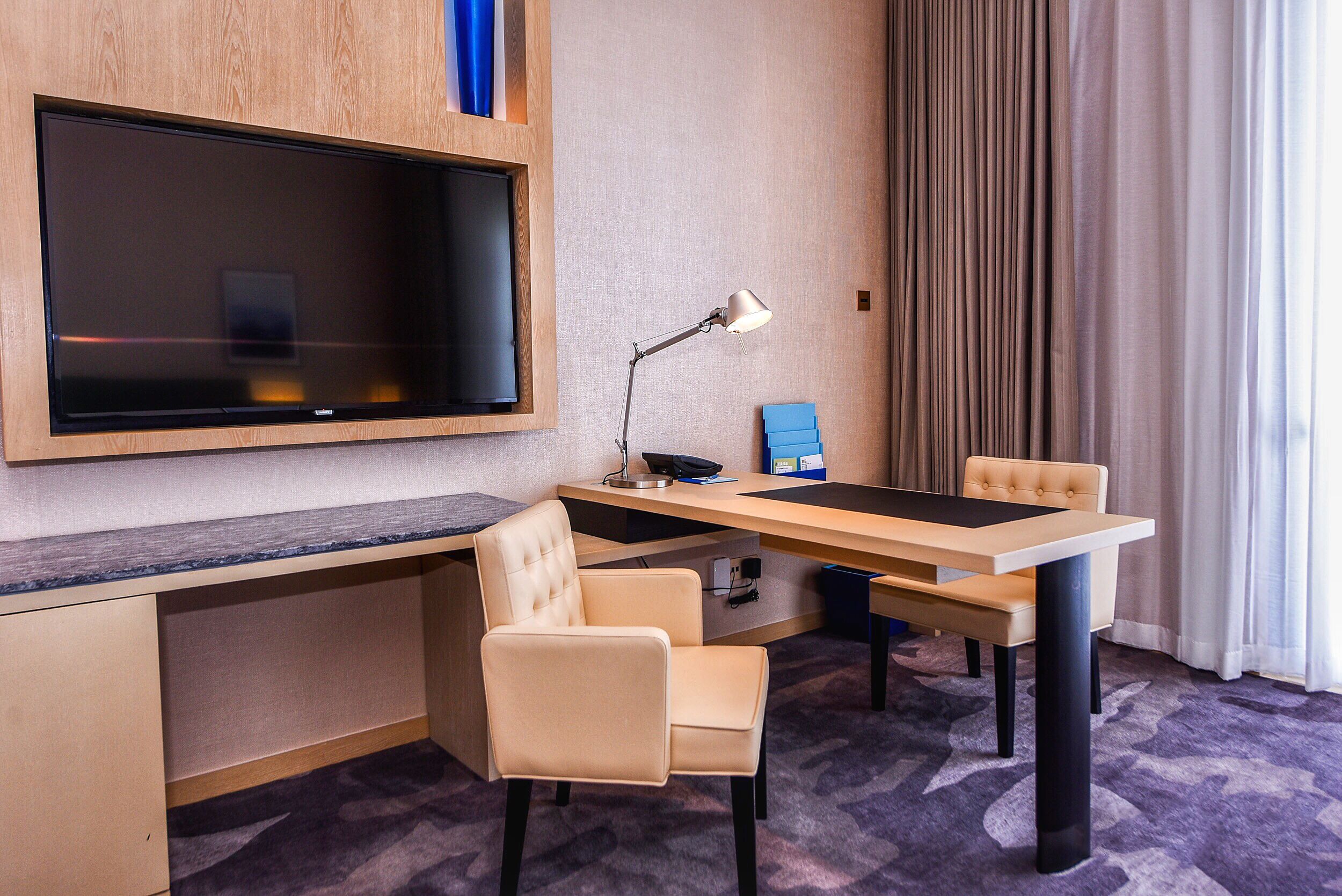 Habitación de hotel con televisión de pantalla plana montada en la pared, un escritorio con una silla, una lámpara y una caja azul sobre el escritorio.