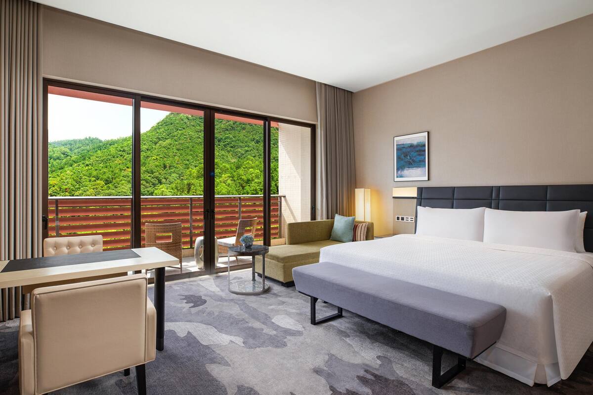 Quarto de hotel com uma cama grande, uma escrivaninha, uma cadeira e uma varanda com vista para uma colina arborizada.