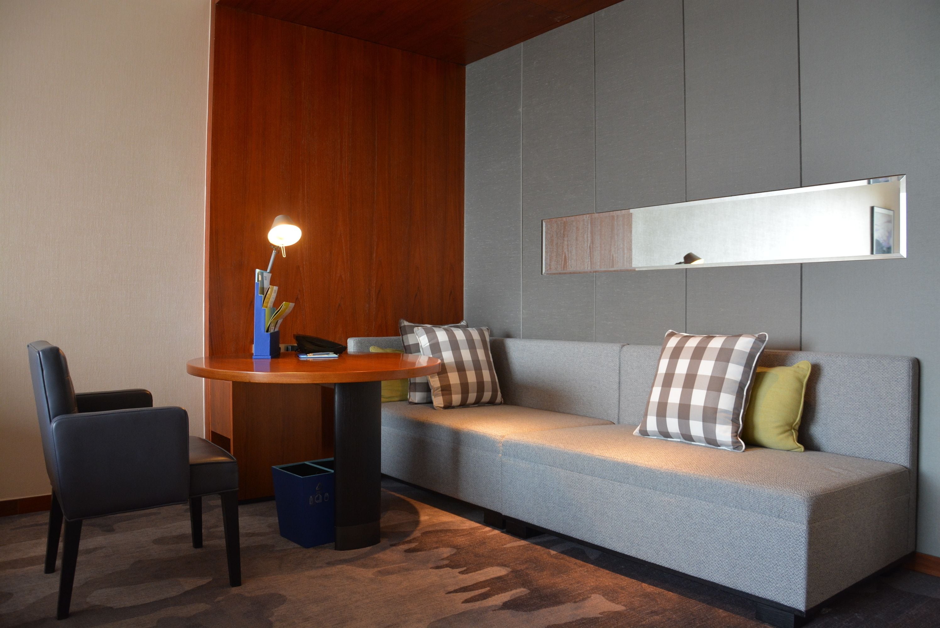Camera d'hotel moderna con un divano grigio, una parete decorata in legno, un tavolino con una lampada e un cestino dei rifiuti blu.