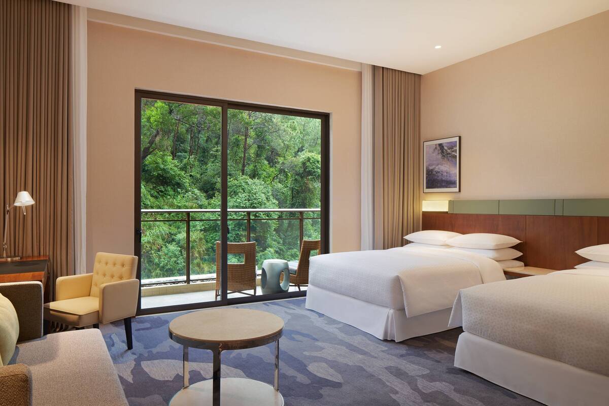 Quarto de hotel com duas camas, um espaço de estar com uma mesa e uma janela grande com vista para o verde.