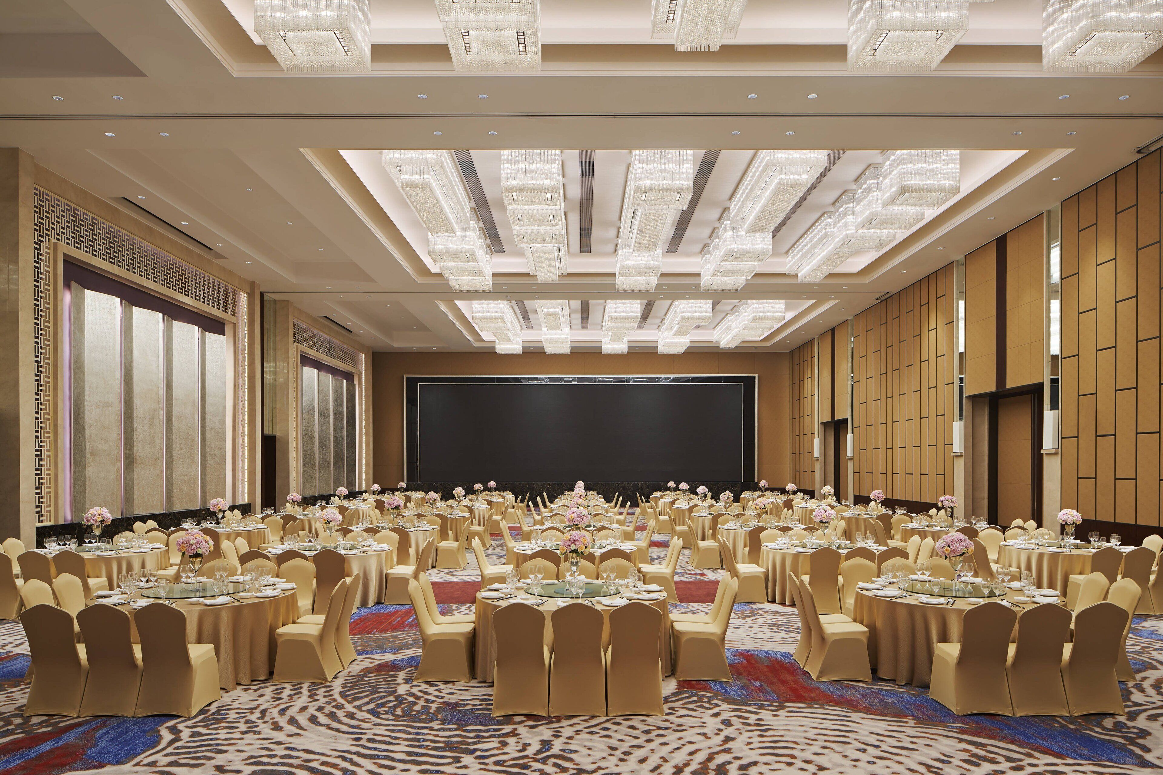 Un salón de conferencias amplio con mesas redondas, sillas color beige y una pantalla grande al frente.